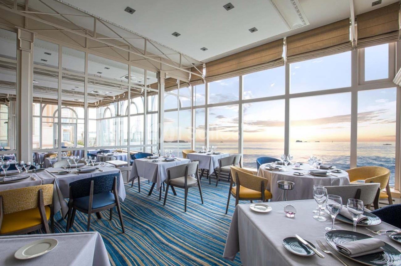Salle de restaurant lumineuse avec grandes baies vitrées offrant une vue sur la mer au coucher du soleil.