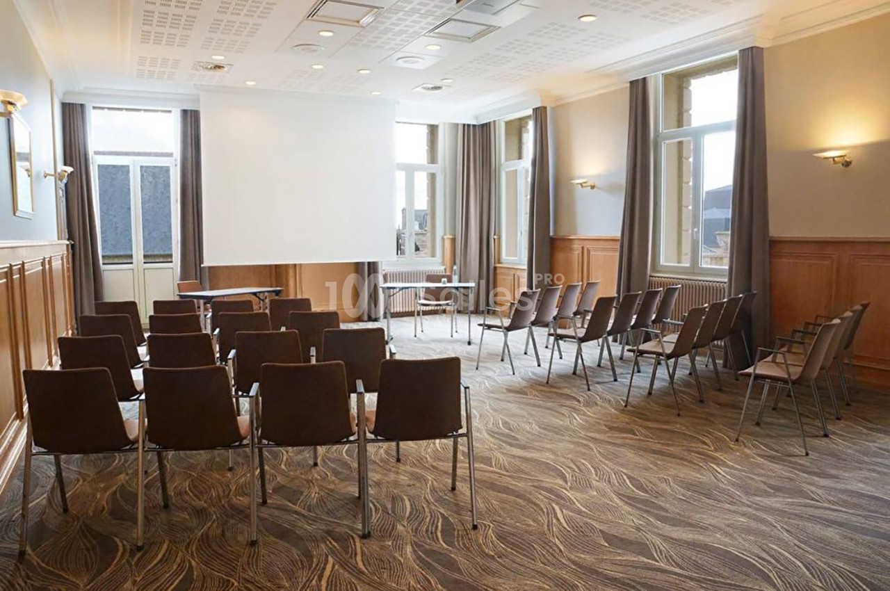Salle de réunion lumineuse avec rangées de chaises, écran de projection et grandes fenêtres donnant sur l'extérieur.