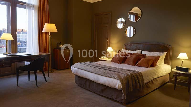 Chambre d'hôtel élégante avec lit double, bureau, lampes allumées et décor chaleureux dans des tons bruns.