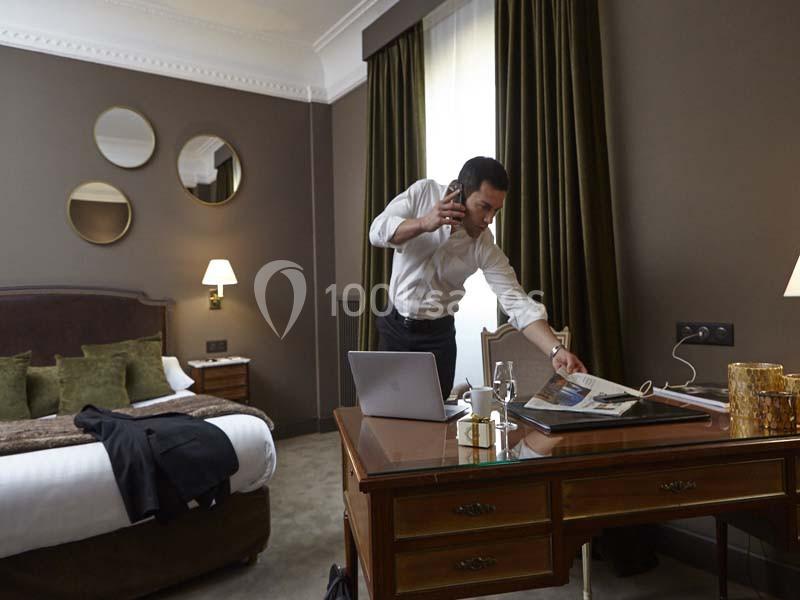 Homme en chemise blanche parlant au téléphone, debout près d'un bureau dans une chambre d'hôtel élégante.