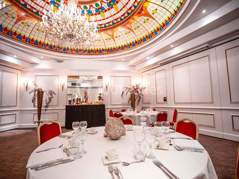 Salle de réception élégante avec tables dressées, chaises rouges et plafond décoré d'un vitrail coloré et d'un lustre.