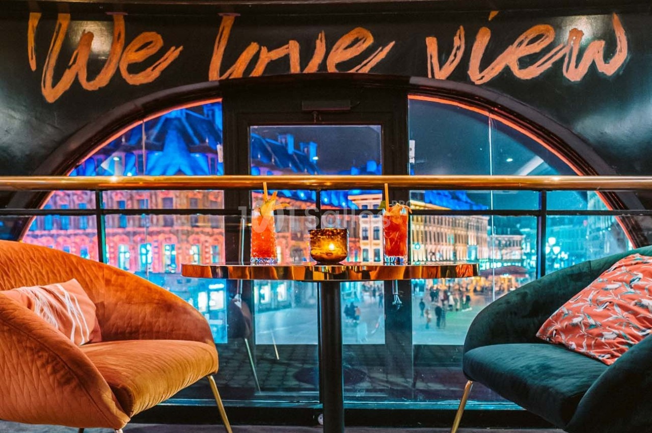 Deux fauteuils colorés autour d'une table avec cocktails, vue nocturne sur une place animée à travers une grande fenêtre.