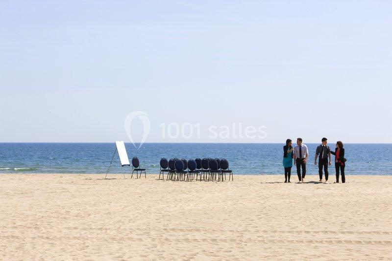 Un groupe de personnes discute sur une plage près de chaises alignées et d'un tableau blanc.