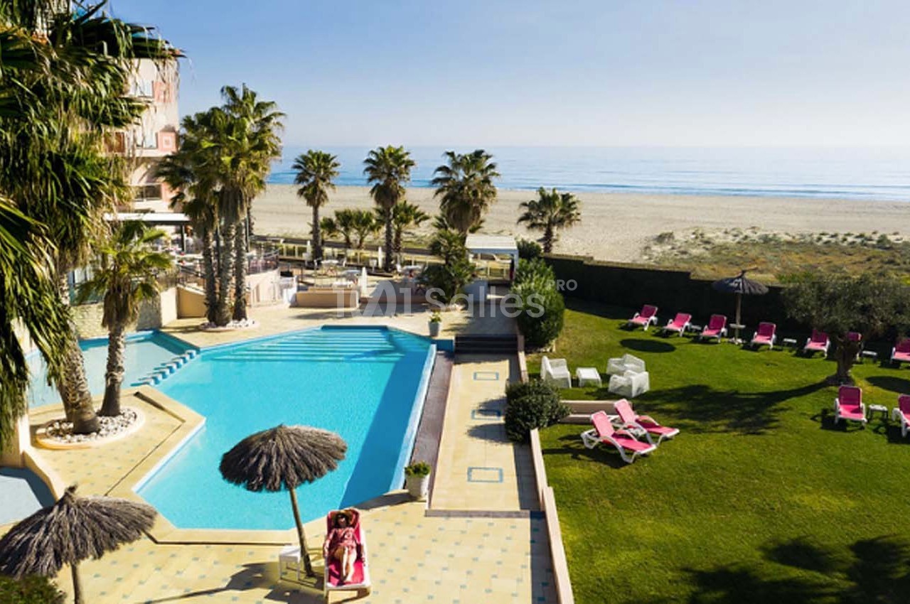 Vue d'une piscine extérieure entourée de palmiers, avec des transats et une plage en arrière-plan.