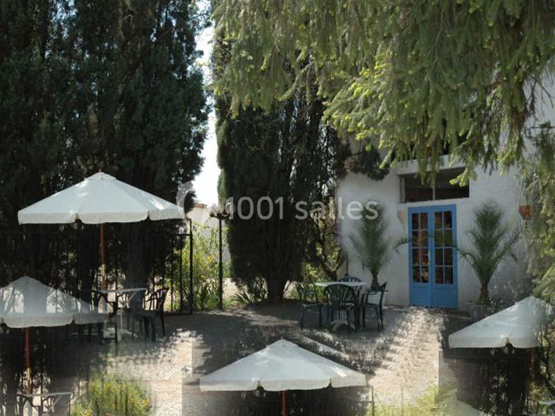Terrasse extérieure avec tables, chaises, parasols blancs, entourée de grands arbres et une porte bleue en arrière-plan.
