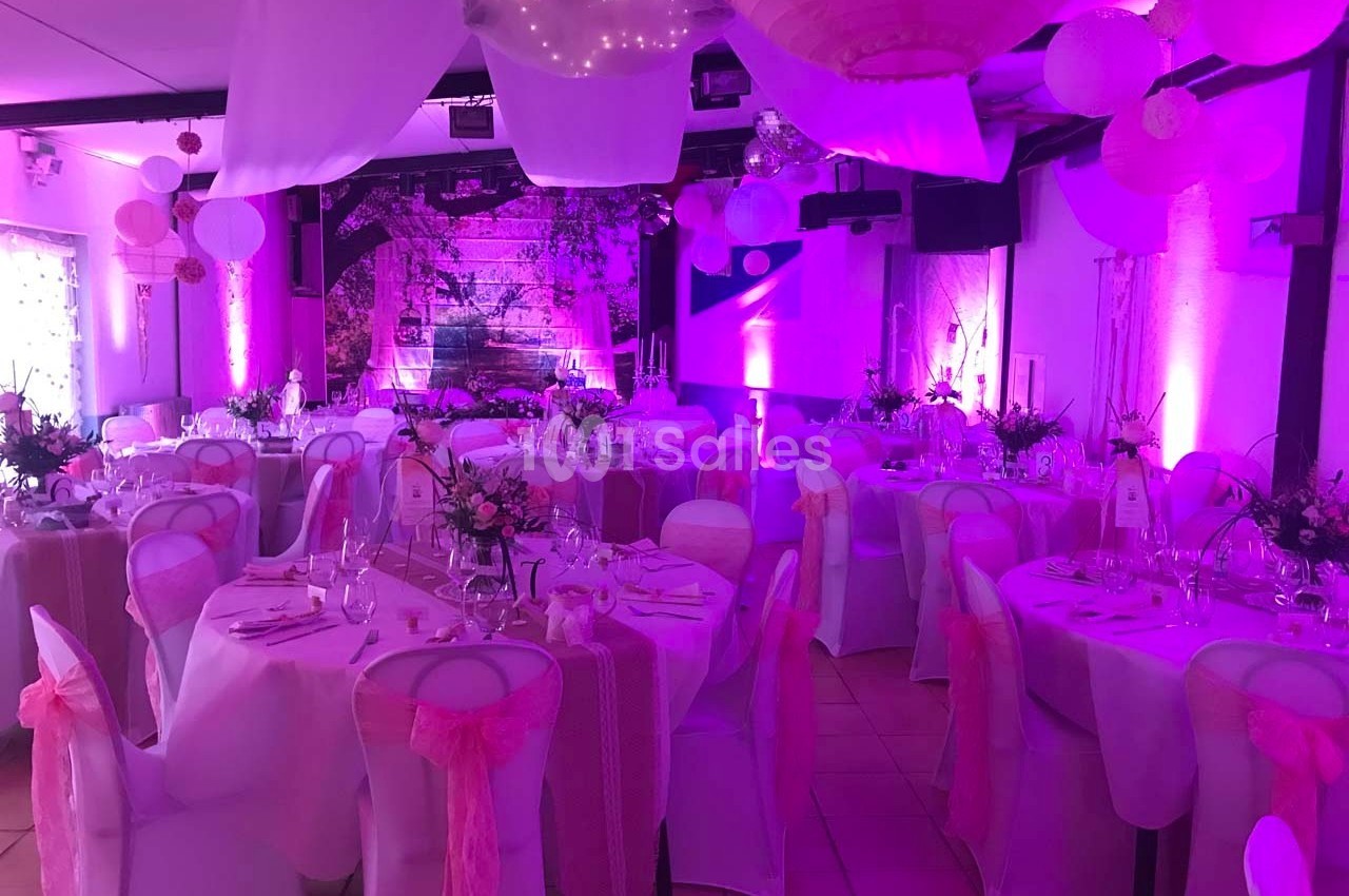 Salle de réception décorée avec des nappes blanches, des rubans roses et un éclairage d'ambiance violet.