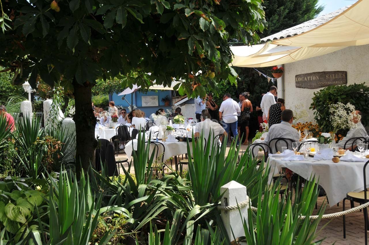 Des convives dînent en plein air dans un jardin ombragé, entourés de végétation et de tables décorées.