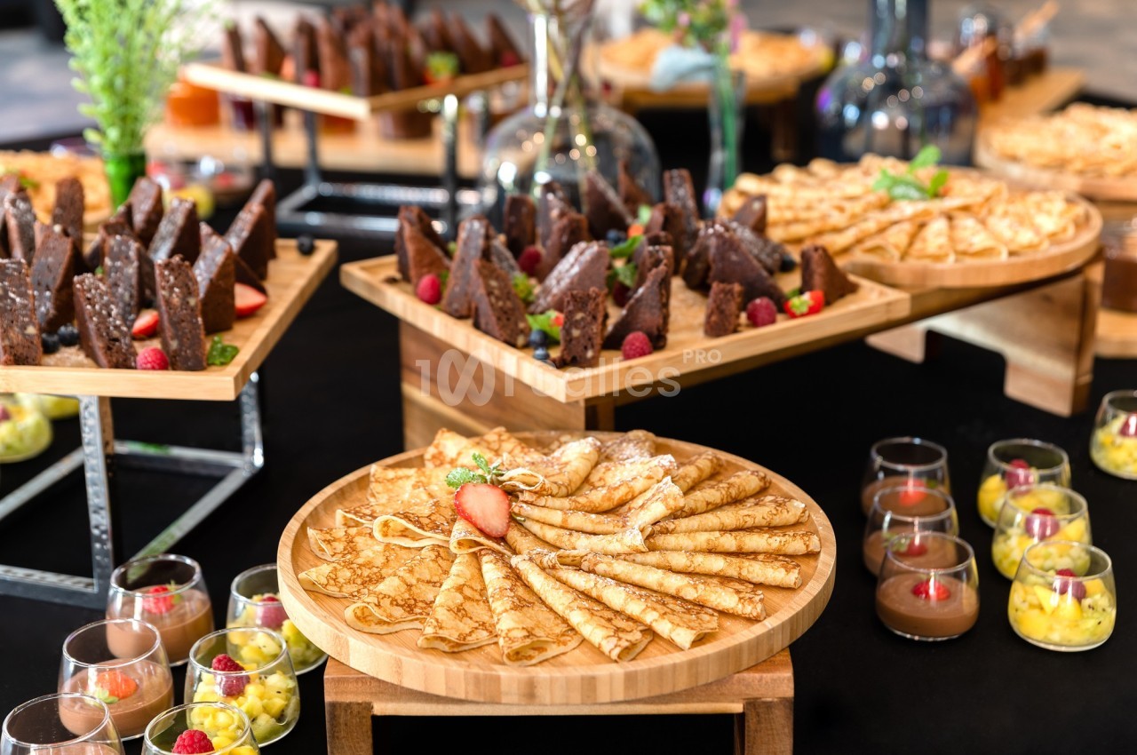 Plateau de crêpes garnies de fruits, entouré de desserts variés comme gâteaux au chocolat et verrines colorées.