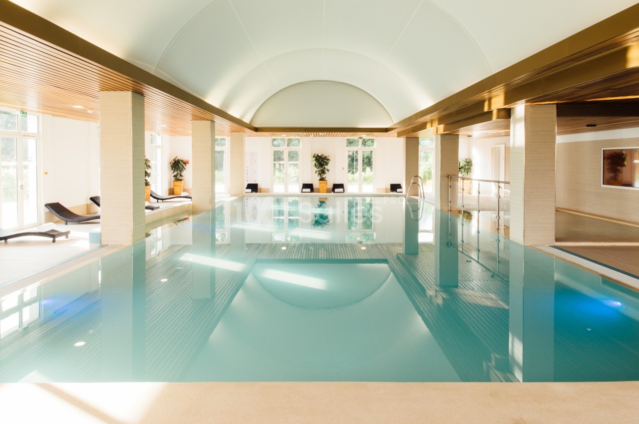 Piscine intérieure lumineuse avec plafond voûté, transats alignés et grandes baies vitrées laissant entrer la lumière…