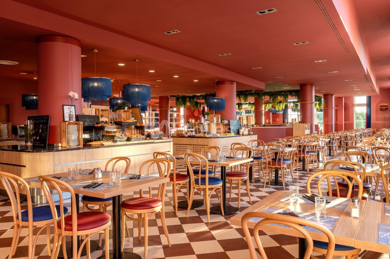 Salle de restaurant lumineuse avec tables en bois, chaises assorties et buffet décoré de plantes suspendues.