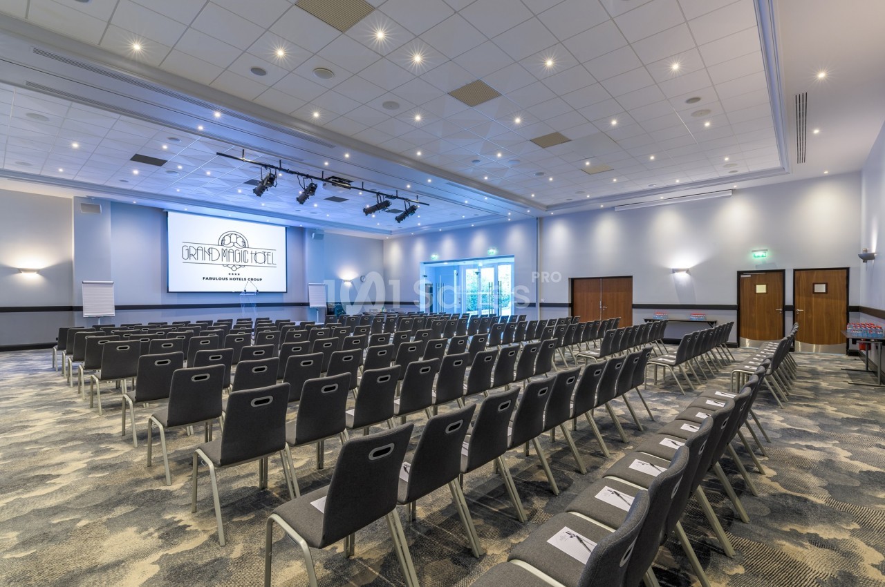 Salle de conférence lumineuse avec rangées de chaises alignées, écran de projection et portes vitrées au fond.