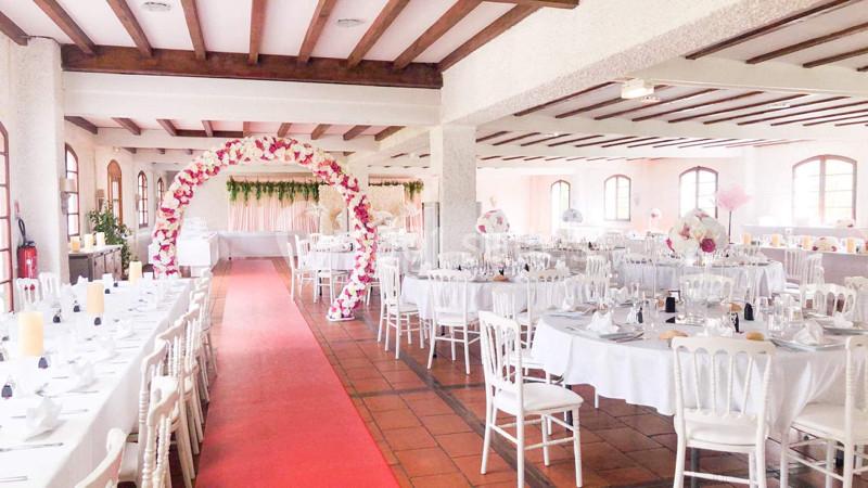 Salle de réception décorée avec des tables dressées, une arche florale et un tapis rouge au centre.