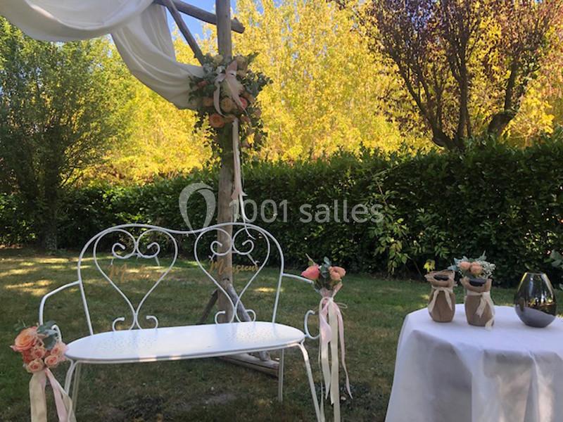 Banc blanc en fer forgé sous une arche décorée de fleurs, avec une table ornée de vases dans un jardin ensoleillé.