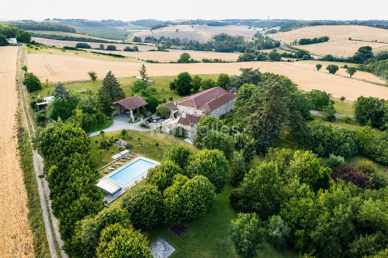 Vue aérienne d'une maison en pierre avec piscine, entourée de jardins verdoyants et de champs vallonnés.