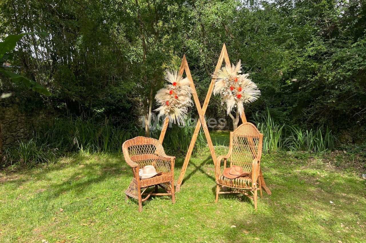 Deux fauteuils en rotin disposés sur une pelouse, encadrés par des structures décorées de fleurs séchées, en extérieur boisé.