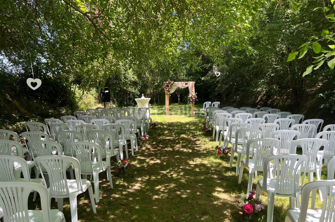 Allée centrale bordée de chaises blanches en plastique, menant à une arche décorée pour une cérémonie en plein air.