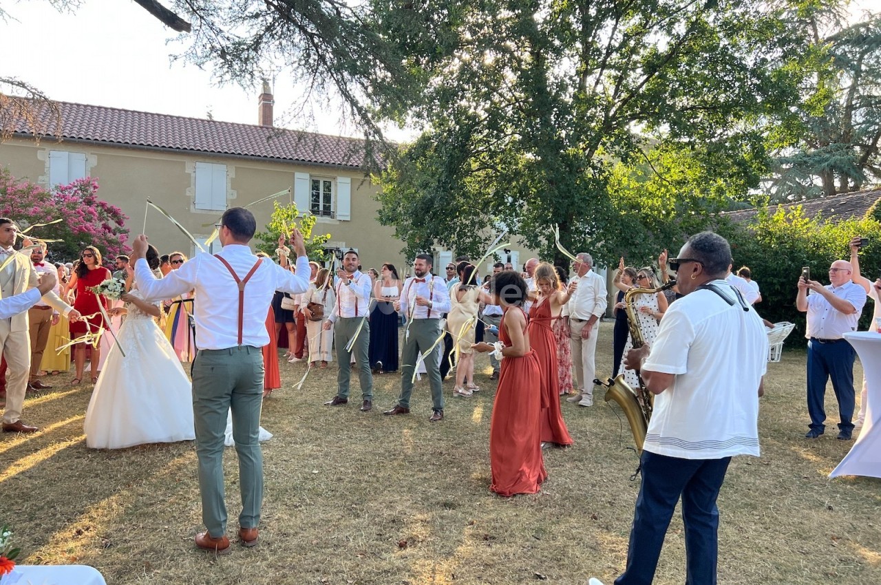 Groupe de personnes célébrant un mariage en extérieur avec des musiciens et des rubans, devant une maison.