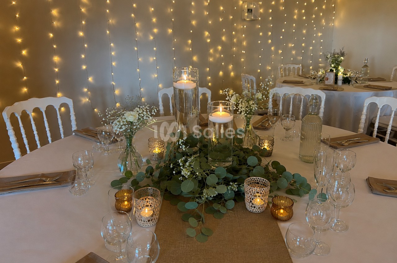 Table décorée pour un événement avec bougies, feuillage, nappes blanches et guirlandes lumineuses en arrière-plan.