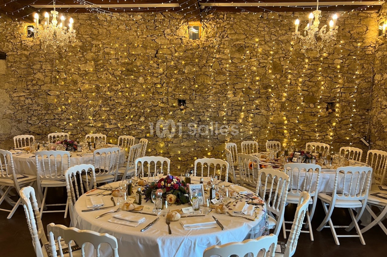 Salle de réception décorée avec des guirlandes lumineuses, des tables rondes dressées et des chaises blanches.