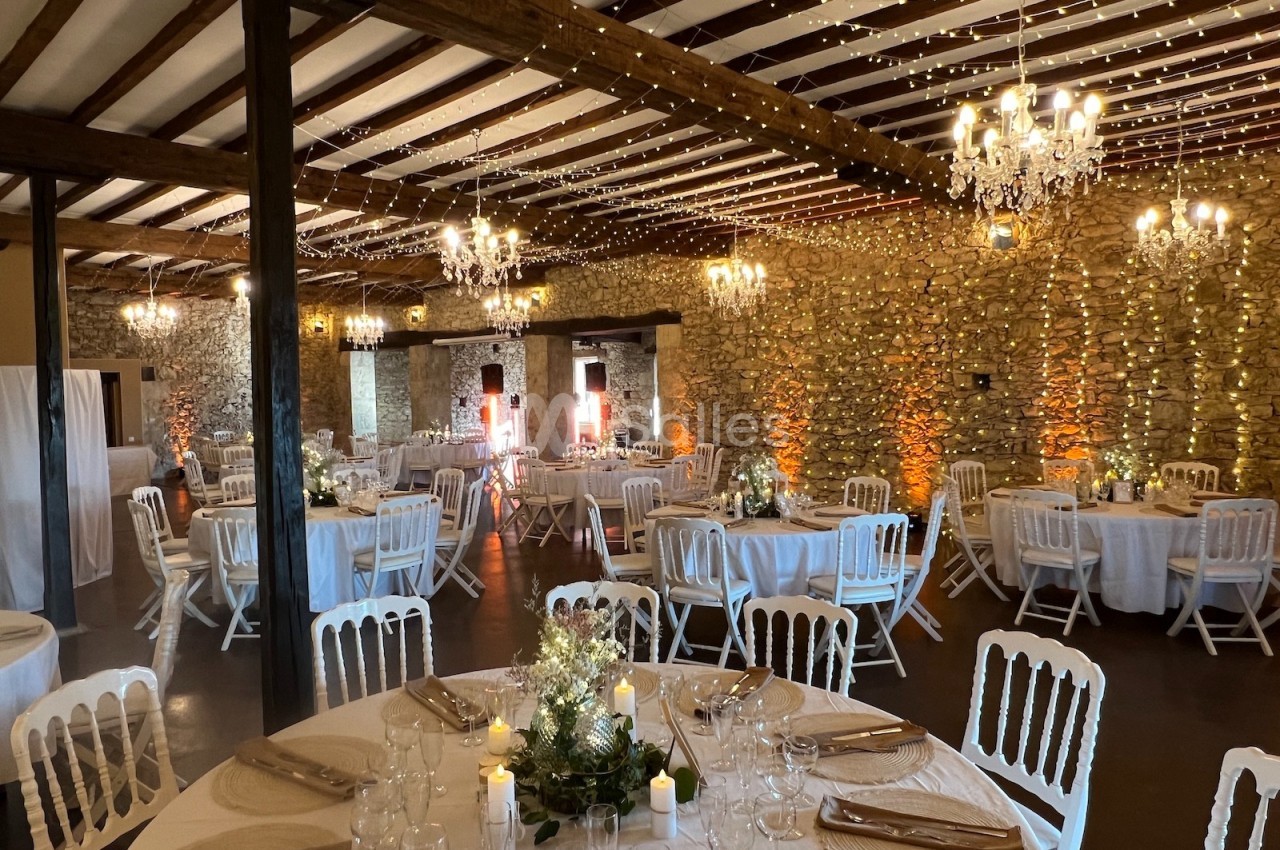 Salle de réception en pierre avec tables rondes dressées, chaises blanches, guirlandes lumineuses et lustres suspendus.