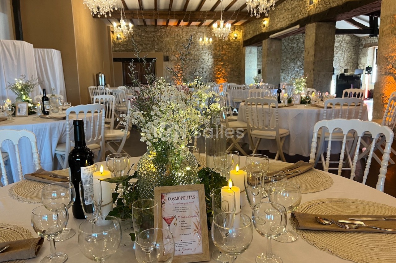 Salle de réception décorée avec des tables rondes, nappes beiges, chandelles, fleurs blanches et chaises blanches.