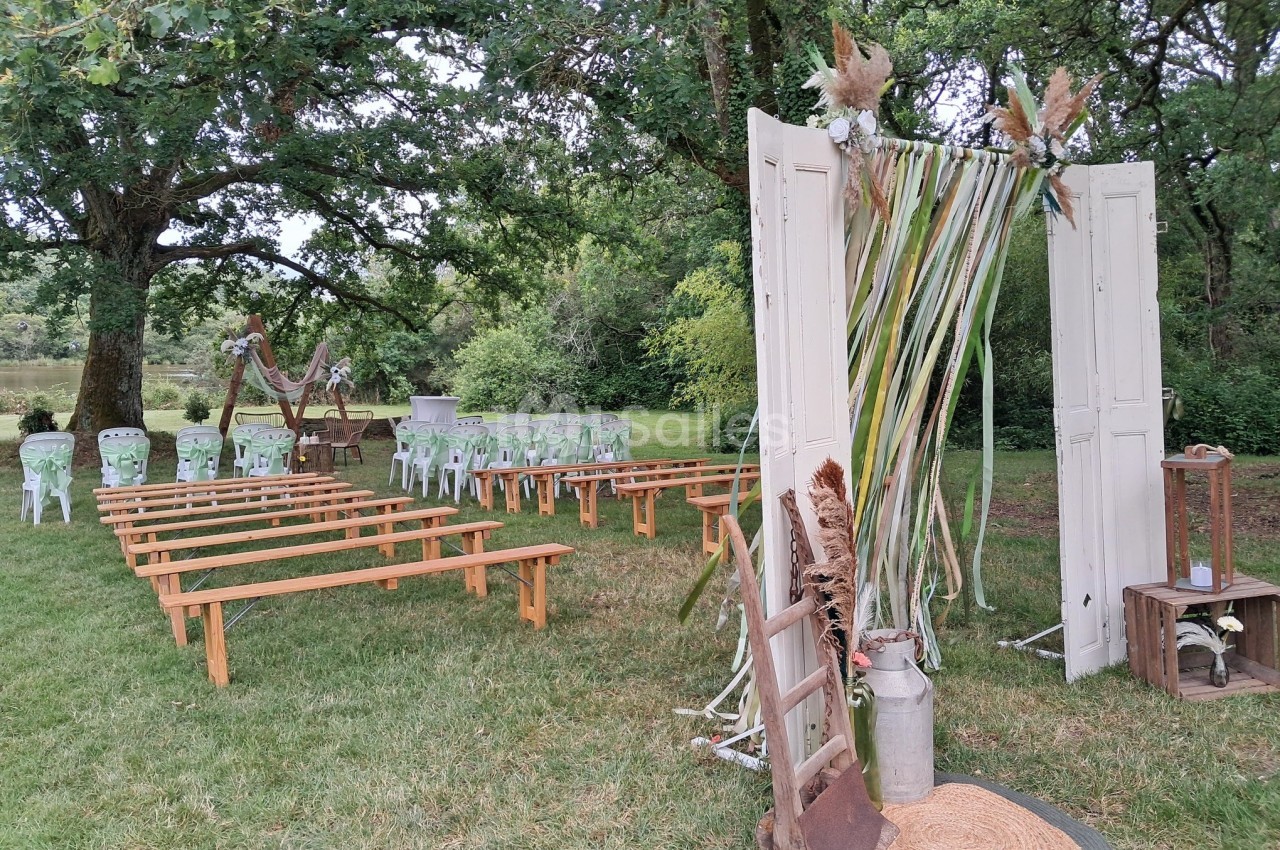 Décoration champêtre pour une cérémonie en plein air avec arche florale, bancs en bois et chaises blanches sur pelouse.