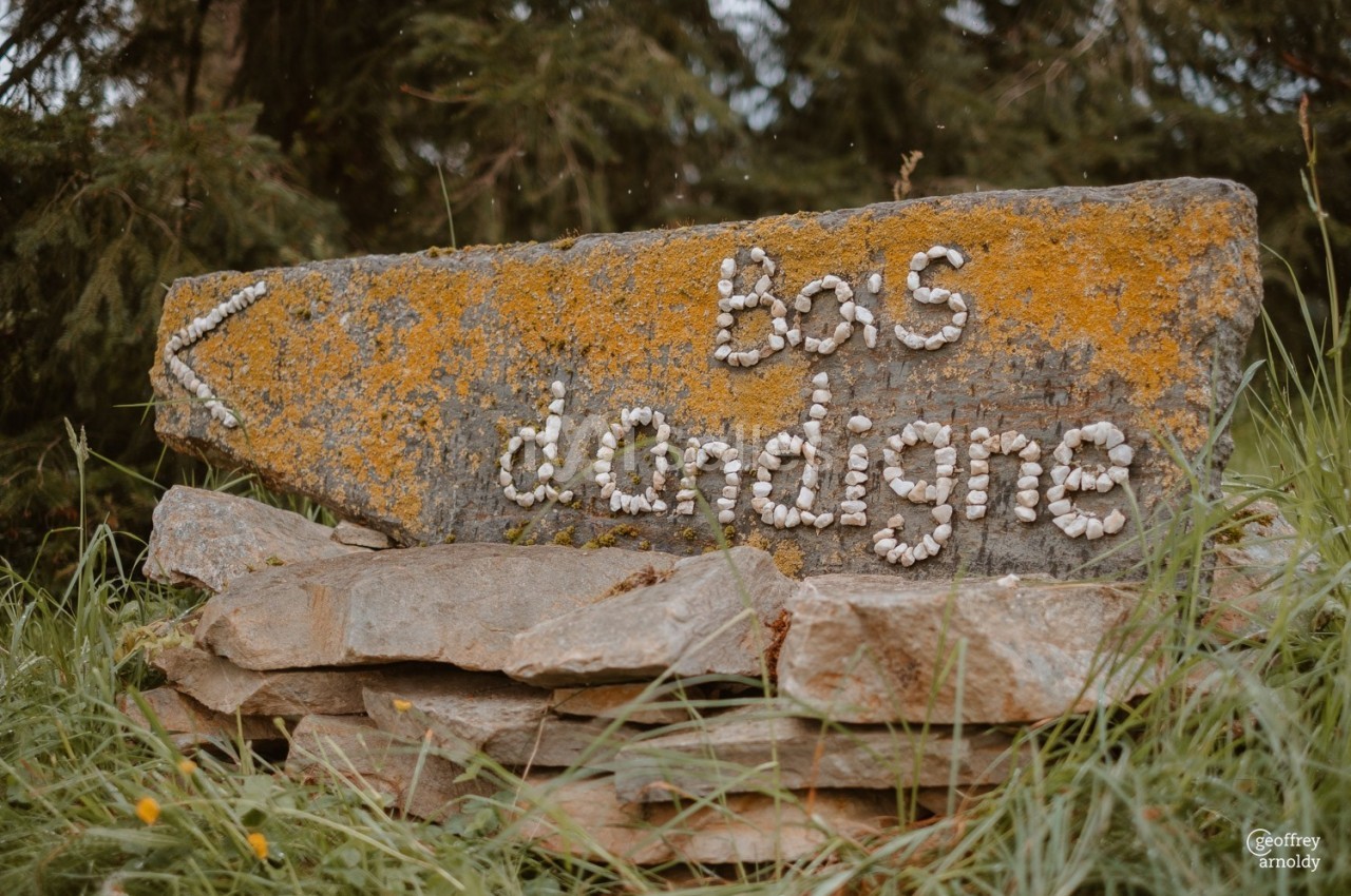Panneau en pierre recouvert de mousse avec l'inscription ’bas d'ardigne’ formée de petits galets, entouré d'herbe.