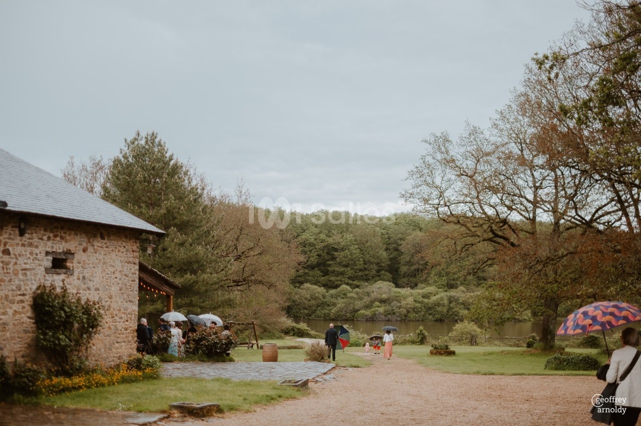 Personnes avec des parapluies marchant près d'un bâtiment en pierre dans un cadre naturel avec arbres et étang.