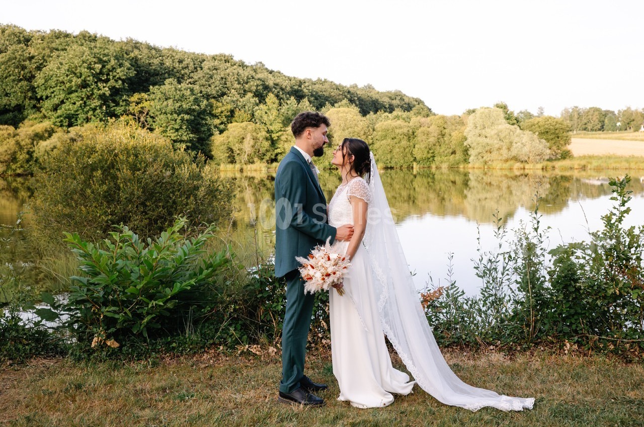 Un couple en tenue de mariage se tient face à face près d'un lac entouré de verdure.