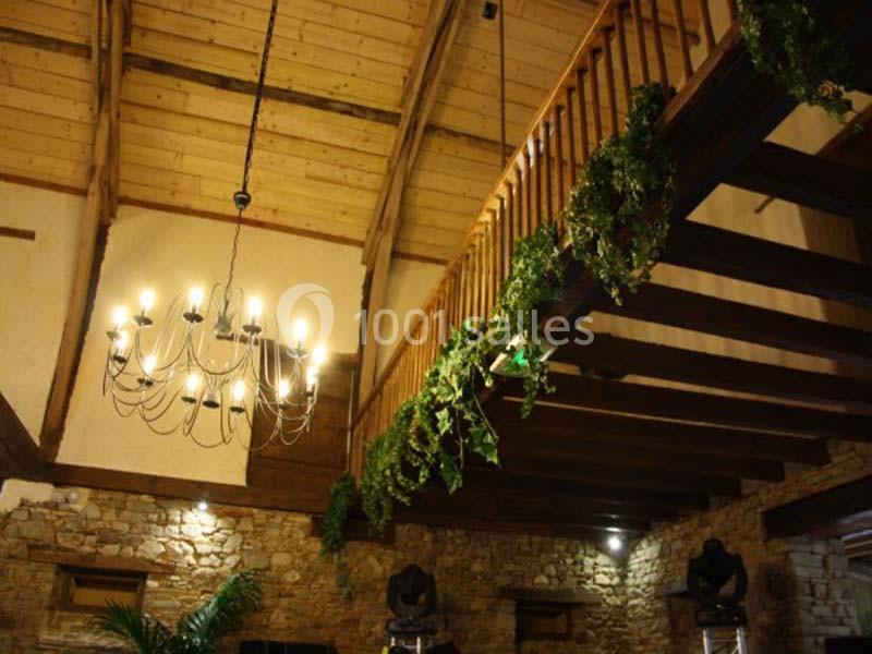 Plafond en bois avec poutres apparentes, lustre suspendu et rambarde décorée de feuillage dans une salle en pierre.