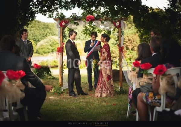 Un mariage en plein air avec un couple devant un officiant sous une arche décorée de fleurs rouges.