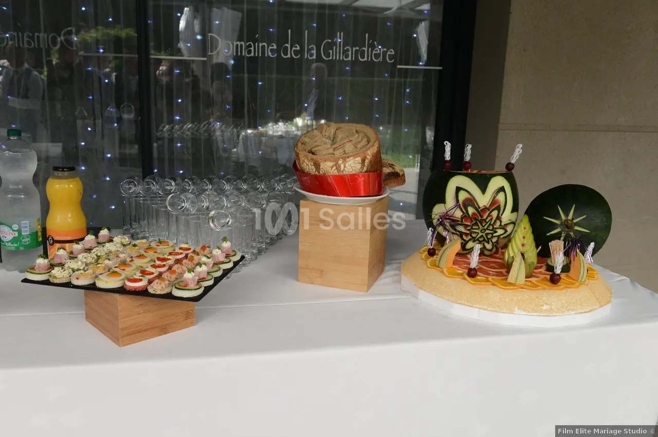 Table dressée avec des amuse-bouches, du pain et des fruits sculptés, devant une baie vitrée décorée de lumières.