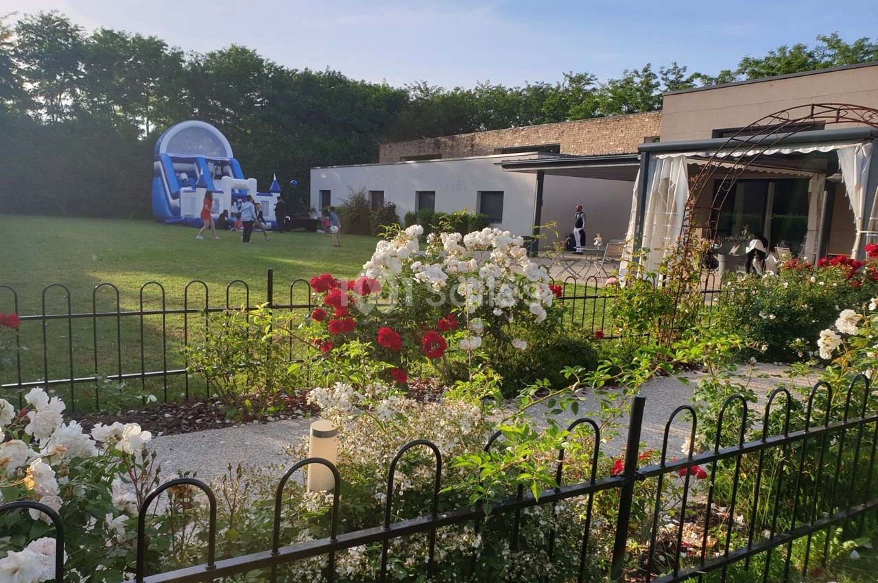 Jardin fleuri avec rosiers, pelouse, structure gonflable en arrière-plan et bâtiment moderne sur la droite.