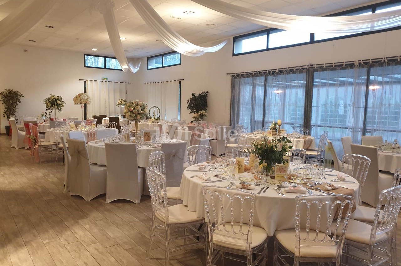 Salle de réception décorée avec des tables rondes dressées, chaises transparentes et voilages suspendus au plafond.