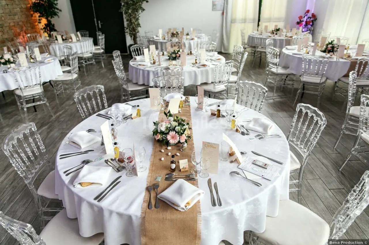 Salle de réception avec tables rondes dressées, nappes blanches, chaises transparentes et décorations florales.