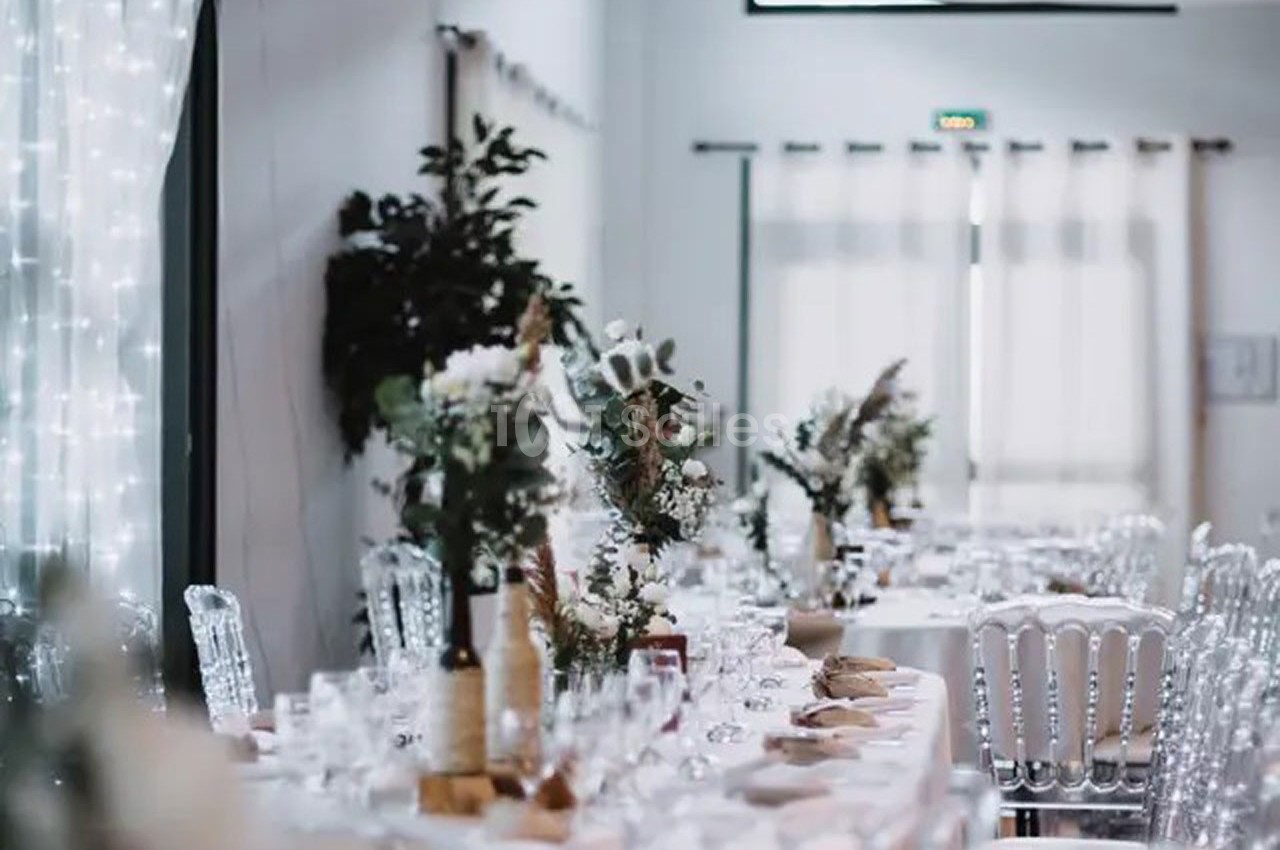 Salle décorée pour un événement avec tables dressées, chaises transparentes et arrangements floraux élégants.