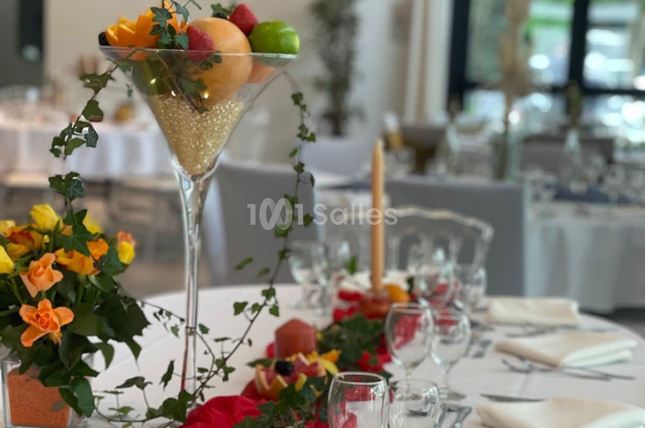 Centre de table avec fruits dans une coupe en verre, fleurs jaunes et rouges, et décoration végétale sur une table dressée.