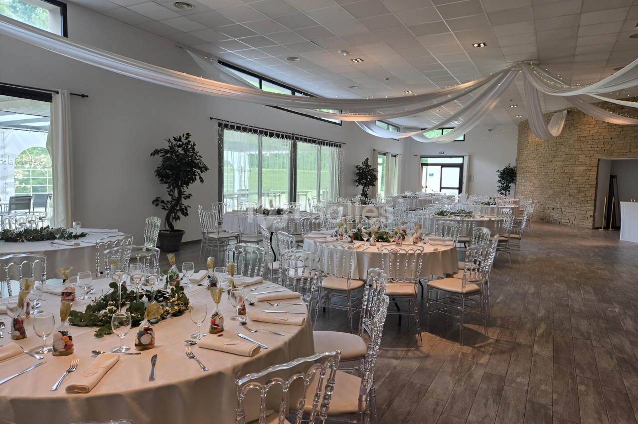 Salle de réception lumineuse avec tables rondes dressées, chaises transparentes et décorations suspendues au plafond.