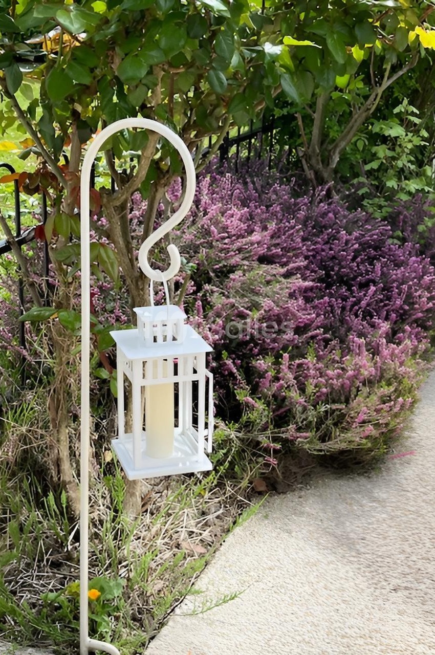 Lanterne blanche suspendue à un crochet métallique devant des buissons de lavande dans un jardin ensoleillé.