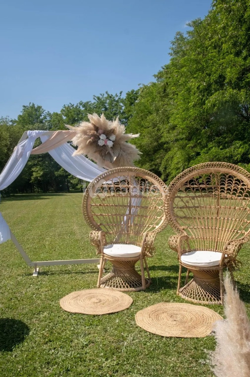 Deux fauteuils en rotin et une arche décorée de fleurs et tissu blanc sur une pelouse entourée d'arbres.