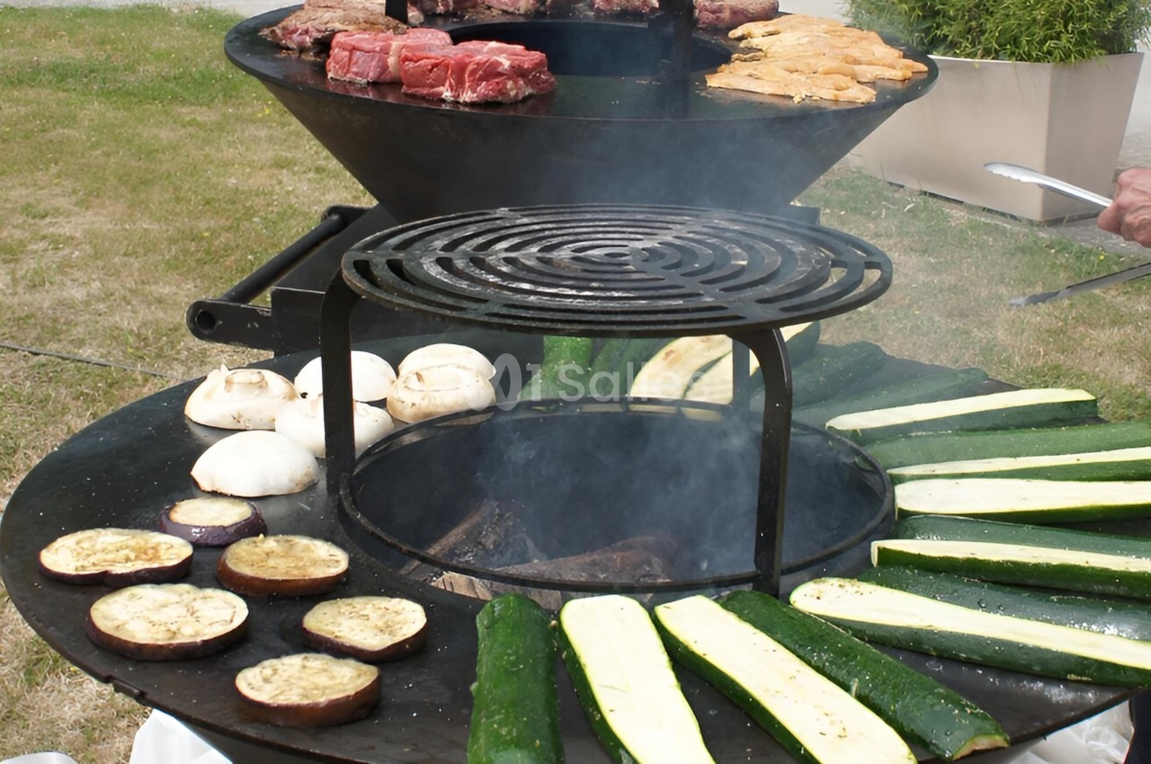 Barbecue circulaire avec légumes grillés (courgettes, aubergines, champignons) et viandes en cuisson sur deux niveaux.