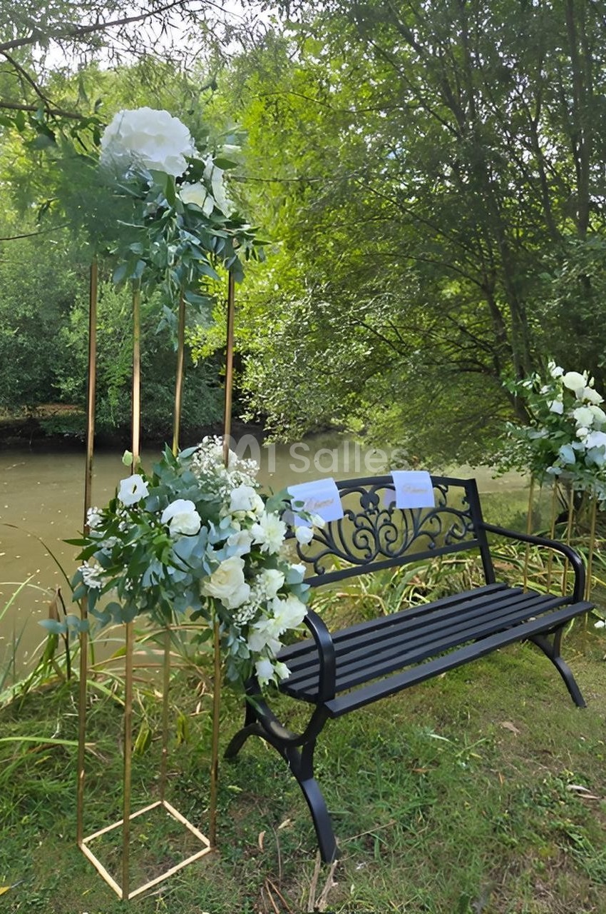 Banc en métal noir décoré de fleurs blanches et feuillages, placé près d'un cours d'eau entouré de verdure.