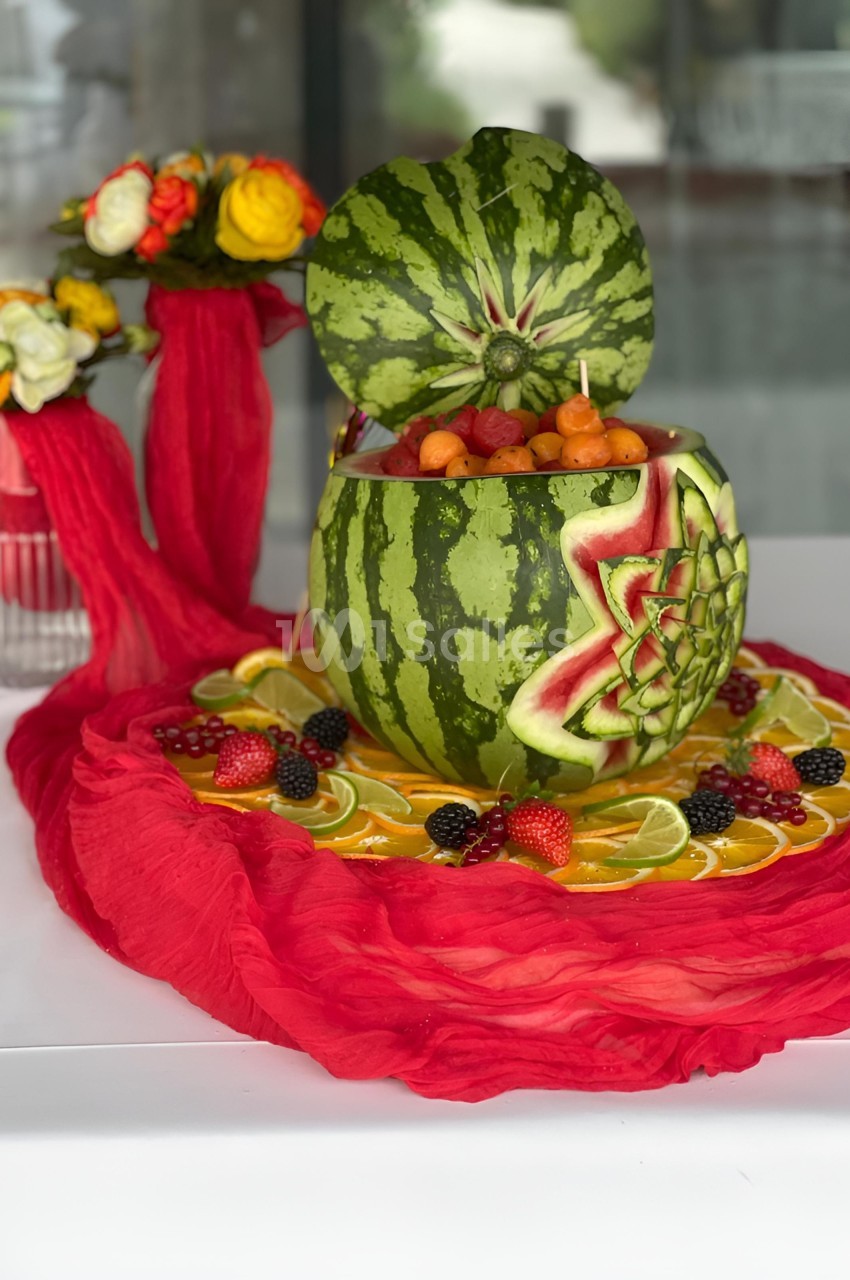 Saladier sculpté dans une pastèque, rempli de fruits variés, entouré de tranches de citron et de fruits rouges.