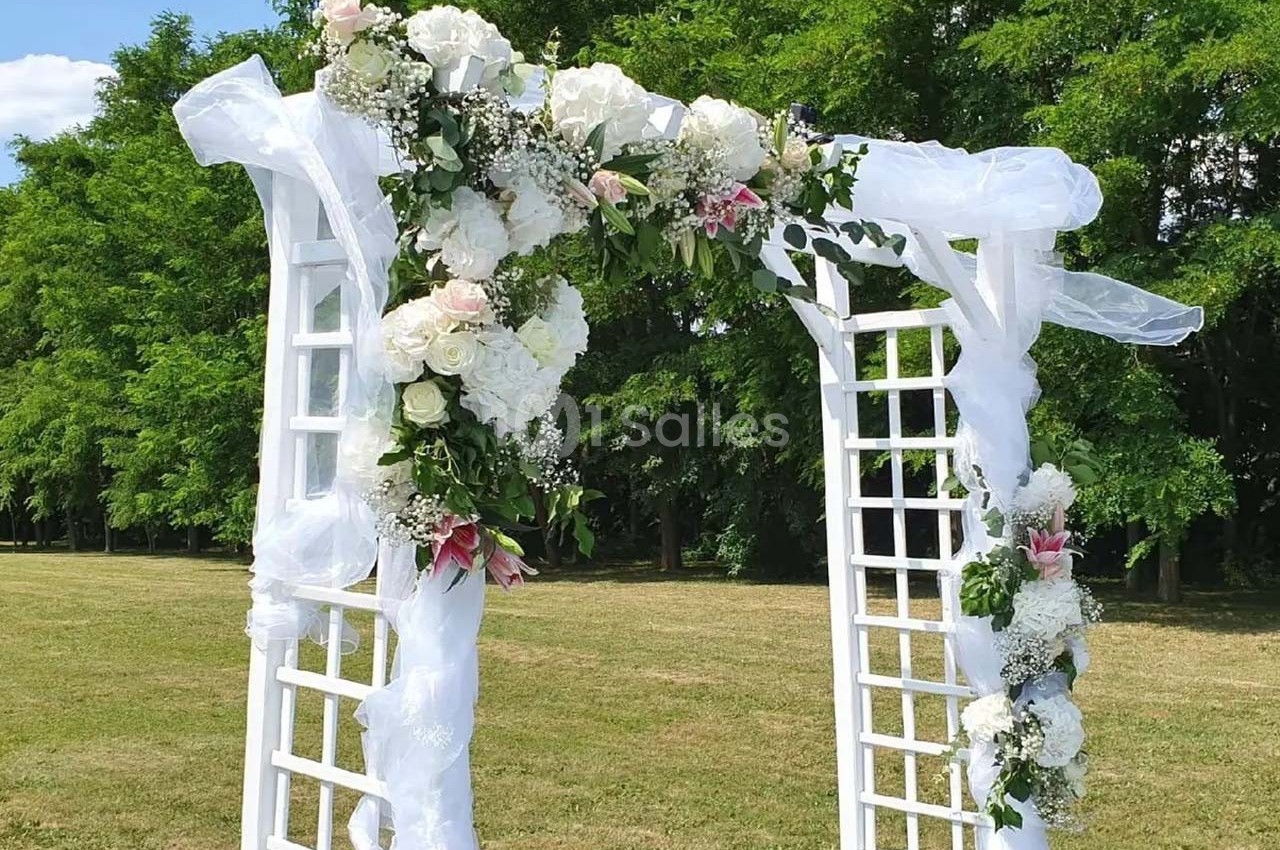 Arche blanche décorée de fleurs blanches et roses avec des voilages, installée dans un parc verdoyant.