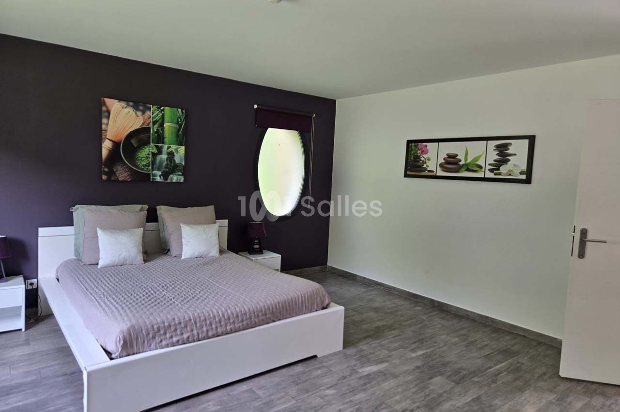 Chambre moderne avec lit double, mur violet décoré de tableaux, sol en parquet gris et fenêtre ovale.