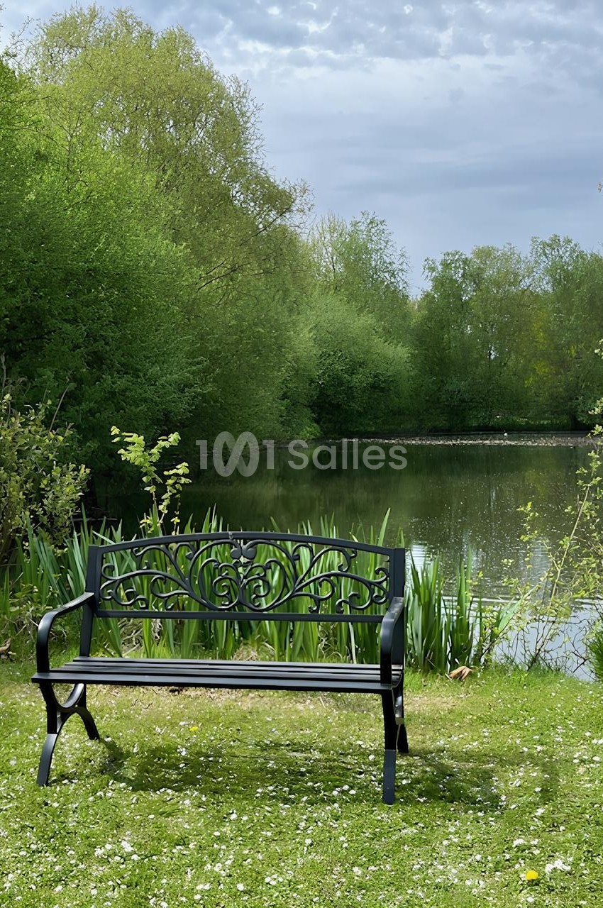 Banc en métal noir placé sur une pelouse, devant un étang entouré de végétation verdoyante sous un ciel nuageux.