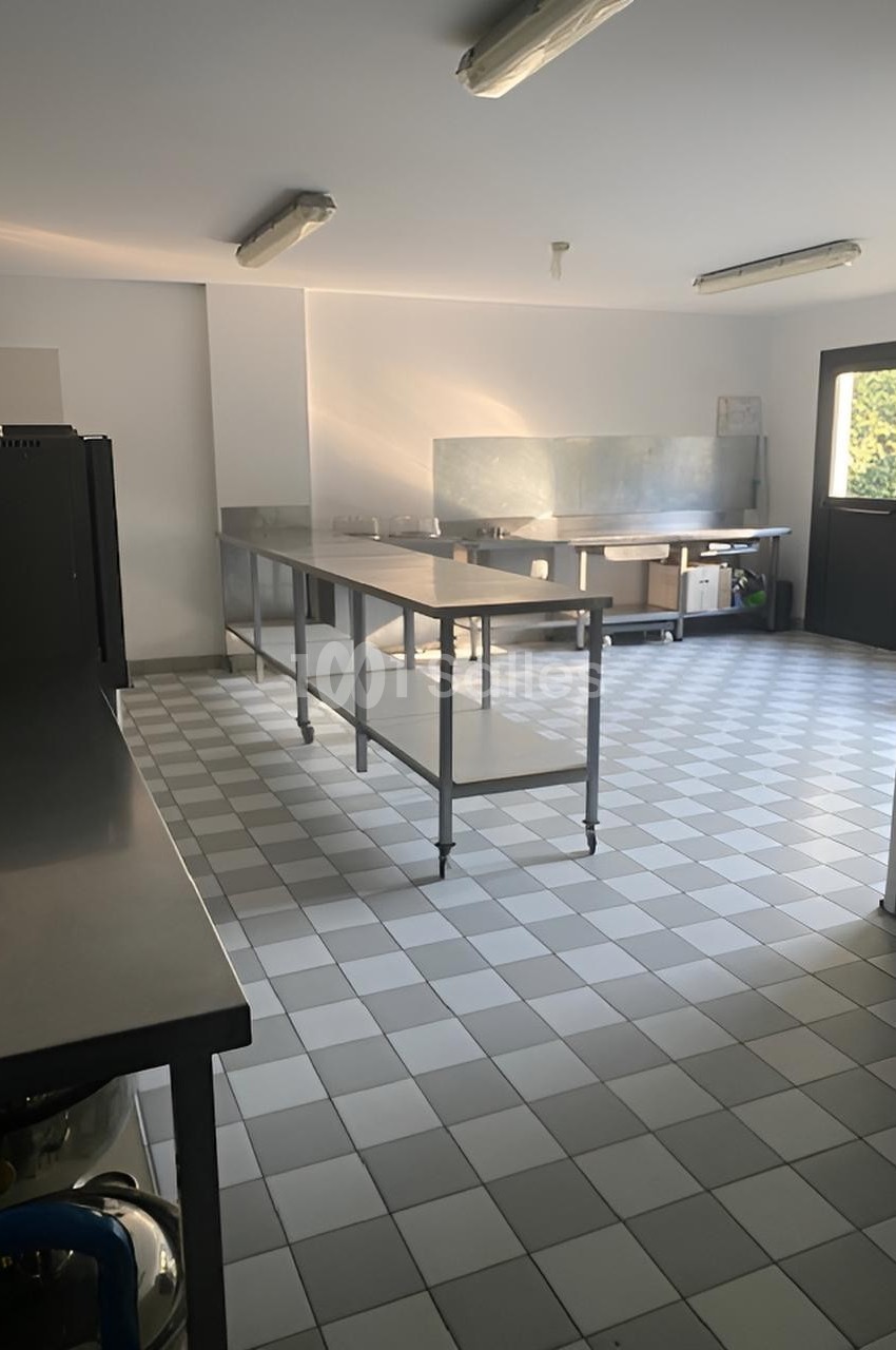 Cuisine professionnelle lumineuse avec plan de travail en inox, sol carrelé et éclairage au plafond.