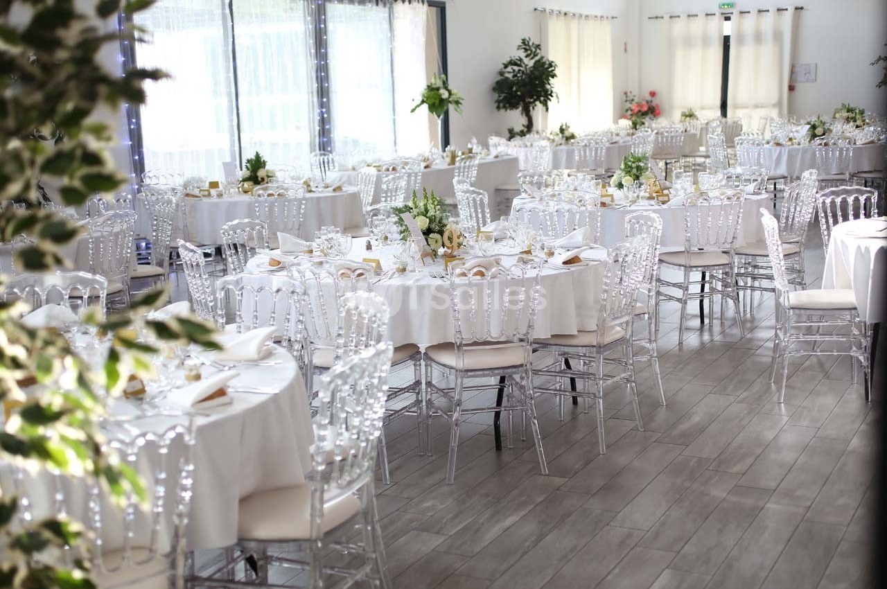 Salle de réception lumineuse avec des tables rondes décorées de nappes blanches, fleurs et chaises transparentes.