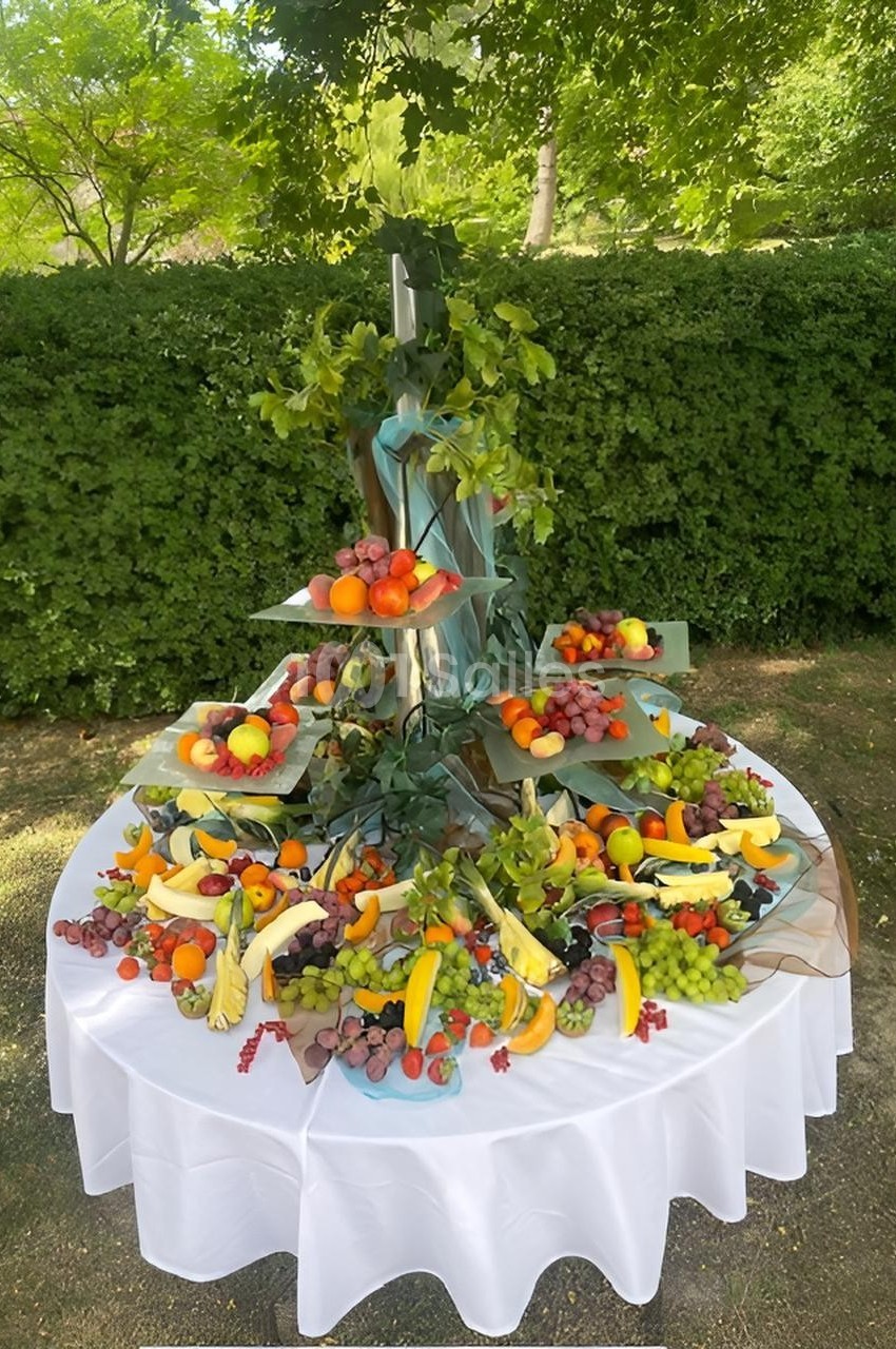 Présentoir de fruits variés disposés sur plusieurs niveaux, entouré de feuillage, sur une table blanche en extérieur.