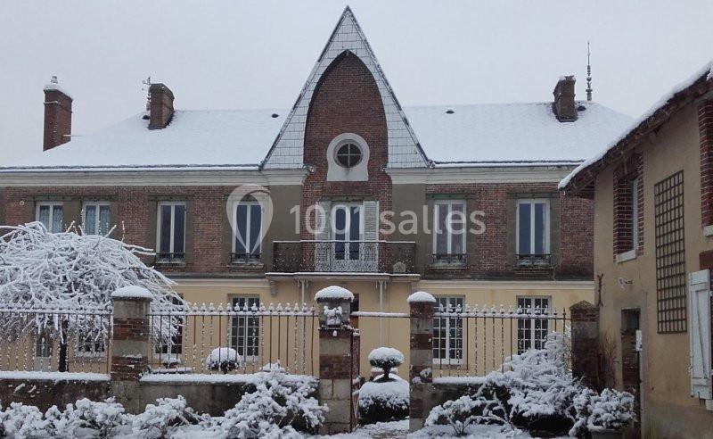 Façade d'une maison en briques avec toit enneigé, jardin et grille en premier plan, sous un ciel gris.